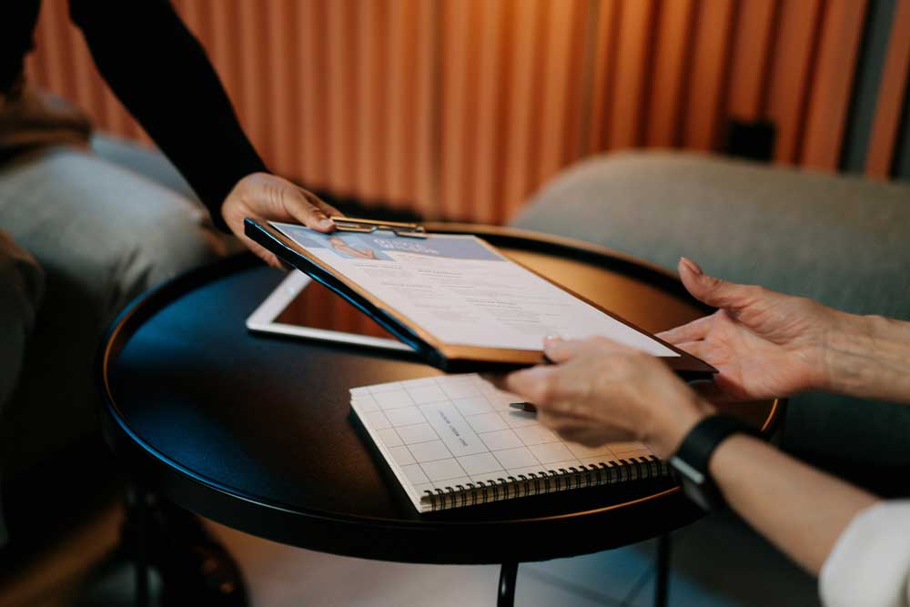 Two individuals reviewing a document on a clipboard, placed on a small round table with a notebook and tablet, symbolizing collaboration and decision-making.