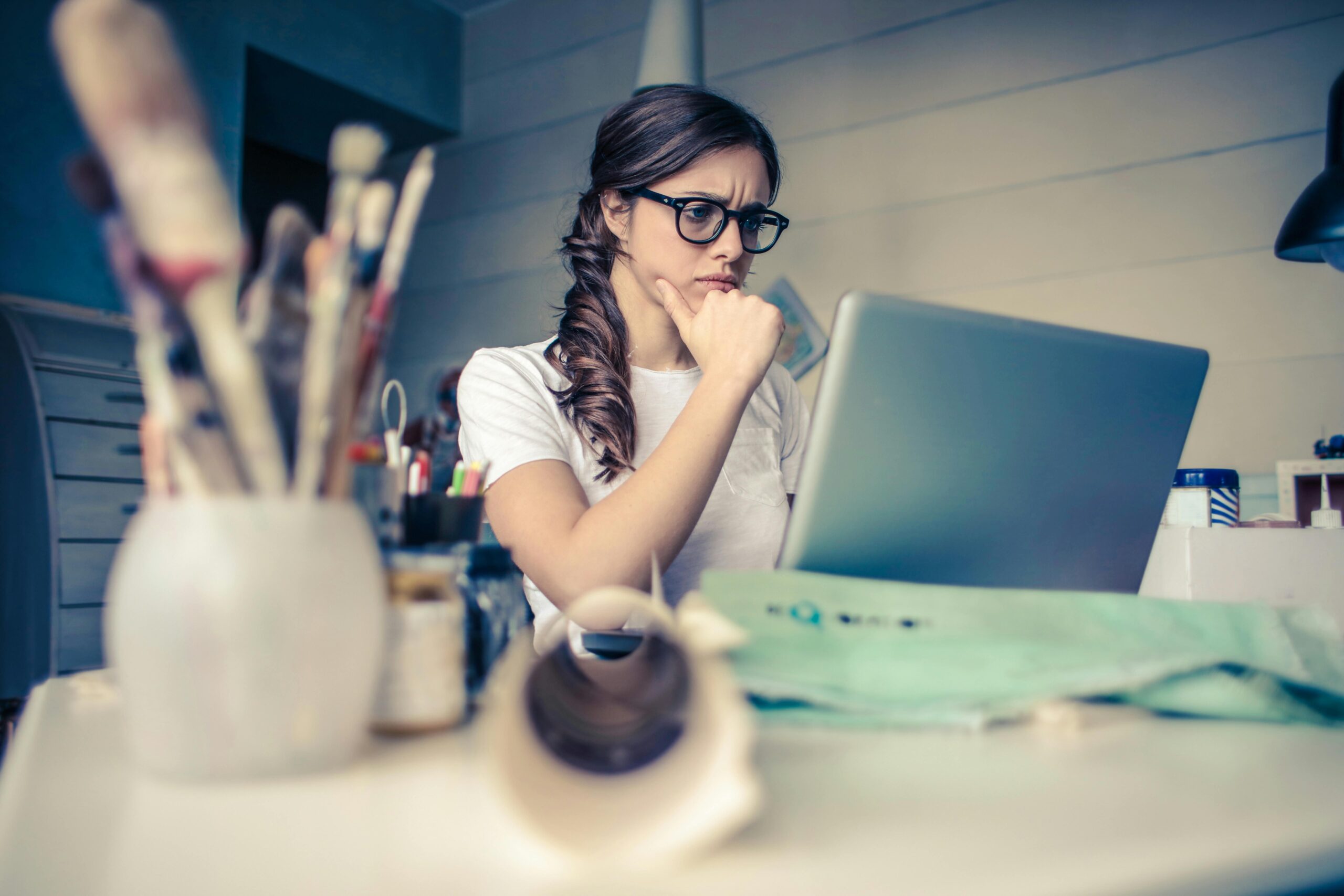 A focused young woman wearing glasses and a white shirt works on her laptop in a creative workspace, surrounded by art supplies. Her expression suggests deep concentration, problem-solving, or analysis.